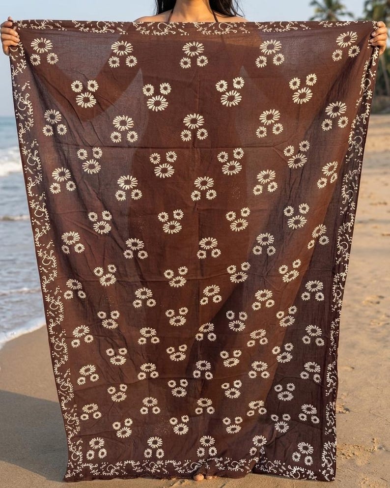 May include: A brown sarong with a white floral pattern and decorative border is held up on a beach. The sarong's design features clusters of white flowers evenly spaced across the fabric. The border has a white, swirling design.