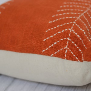 May include: An orange decorative pillow with a white leaf design stitched on the top. The bottom of the pillow is a cream color. The pillow is square and appears to be made of a textured fabric.