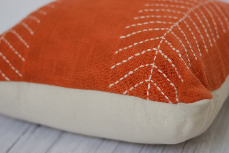 May include: An orange decorative pillow with a white leaf design stitched on the top. The bottom of the pillow is a cream color. The pillow is square and appears to be made of a textured fabric.
