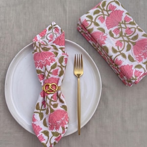 Puede incluir: Una servilleta blanca doblada con un estampado floral rosa y un servilletero dorado se encuentra en un plato blanco junto a un tenedor dorado. Una pila de servilletas a juego está a la derecha. Las servilletas tienen un diseño floral rosa con hojas verdes.