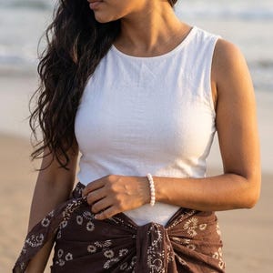 May include: A white sleeveless top and a brown sarong with a floral pattern are worn. A white beaded bracelet is on the wrist. The background is a beach scene with the ocean and sand.