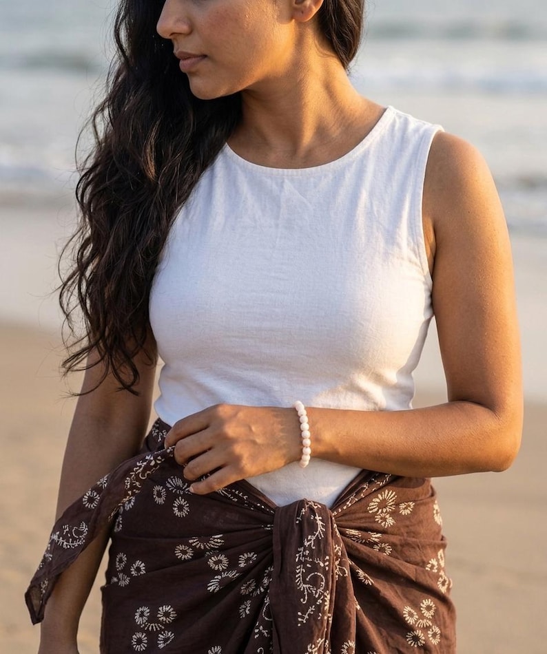 May include: A white sleeveless top and a brown sarong with a floral pattern are worn. A white beaded bracelet is on the wrist. The background is a beach scene with the ocean and sand.