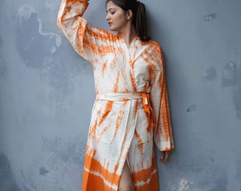 May include: A white and orange tie-dye robe with long sleeves and a matching belt. The robe has a wrap-around design and pockets. The model is wearing the robe against a blue-gray wall.