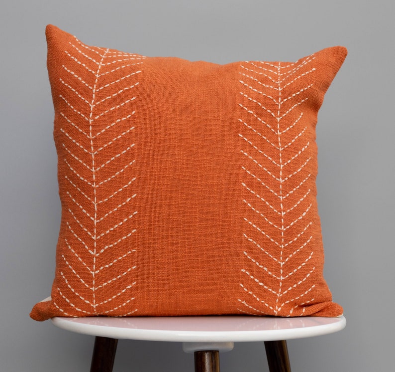 May include: Square orange decorative pillow with white stitched chevron pattern. The pillow is made of textured fabric and is displayed on a white stool. The pillow is a home decor item.