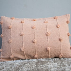 May include: A rectangular peach-colored decorative pillow with vertical lines of textured, knotted accents. The pillow is made of a woven fabric and rests on a light-colored, textured surface. The pillow's design adds a touch of texture and visual interest.