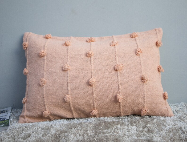 May include: A rectangular peach-colored decorative pillow with vertical lines of textured, knotted accents. The pillow is made of a woven fabric and rests on a light-colored, textured surface. The pillow's design adds a touch of texture and visual interest.