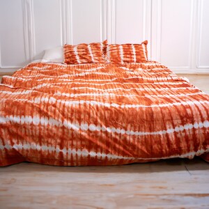 May include: A bed with an orange and white tie-dye patterned duvet cover and two matching pillowcases. The bedding is on a bed with a white pillow and is set against a white paneled wall and a light wood floor.
