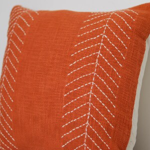 May include: An orange decorative pillow with white stitched chevron patterns. The pillow has a textured fabric and a neutral-colored backing. This accent pillow is suitable for a living room or bedroom.