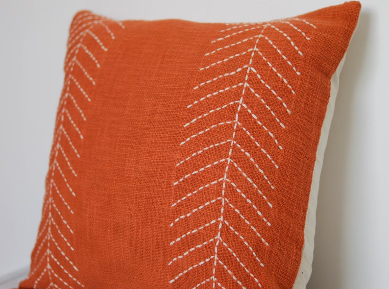 May include: An orange decorative pillow with white stitched chevron patterns. The pillow has a textured fabric and a neutral-colored backing. This accent pillow is suitable for a living room or bedroom.