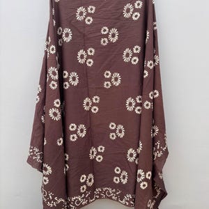 May include: A brown shawl with a repeating pattern of white floral designs. The shawl is draped, showcasing its fabric and the detailed floral pattern. The edges feature a decorative white border.