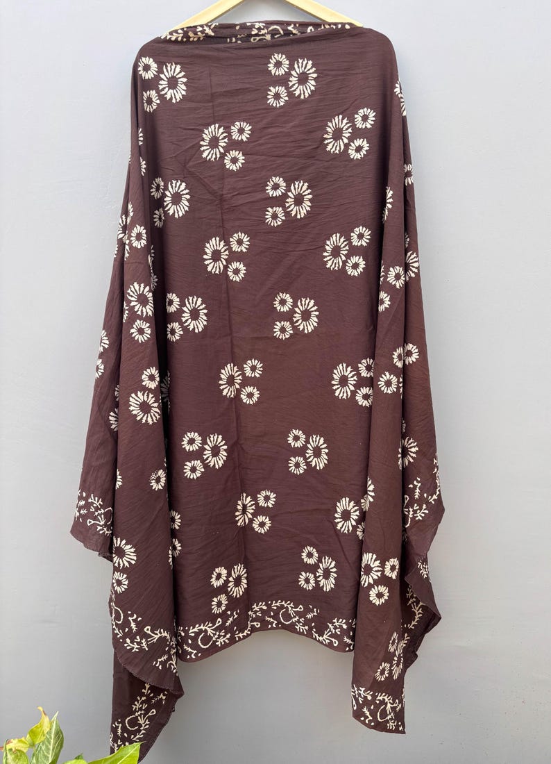 May include: A brown shawl with a repeating pattern of white floral designs. The shawl is draped, showcasing its fabric and the detailed floral pattern. The edges feature a decorative white border.