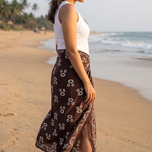 May include: A brown sarong with a white floral pattern. The sarong is worn over a white tank top. The image is taken on a beach with the ocean in the background. The sarong appears to be made of a lightweight fabric.