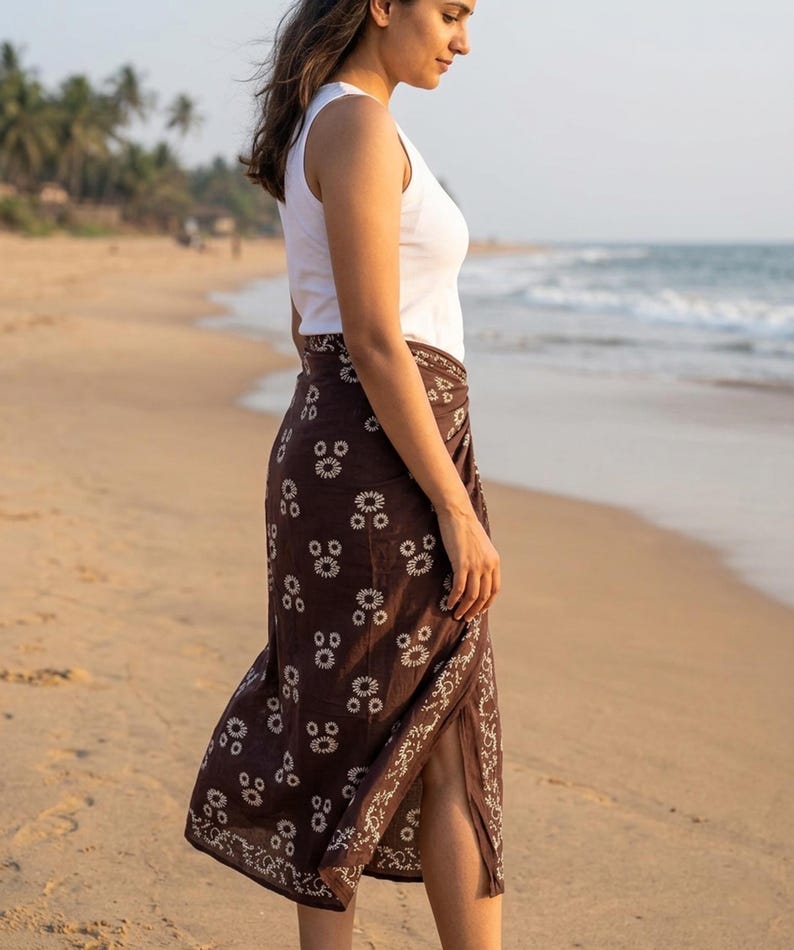 May include: A brown sarong with a white floral pattern. The sarong is worn over a white tank top. The image is taken on a beach with the ocean in the background. The sarong appears to be made of a lightweight fabric.