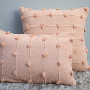 May include: Two peach-colored decorative pillows. The pillows have a textured fabric with vertical lines and small, round, tufted accents. One pillow is rectangular, and the other is square.