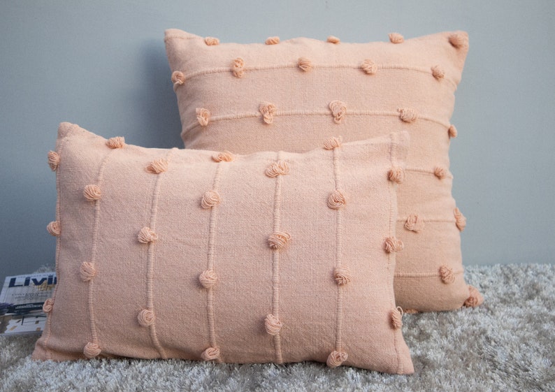 May include: Two peach-colored decorative pillows. The pillows have a textured fabric with vertical lines and small, round, tufted accents. One pillow is rectangular, and the other is square.