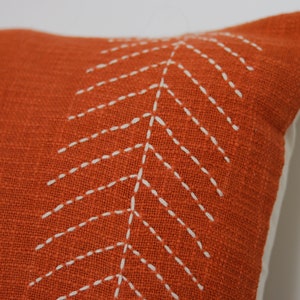 May include: Close-up of an orange decorative pillow with a white stitched leaf design. The pillow has a textured fabric and a white backing. The pillow is a home decor item.