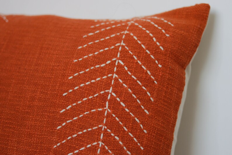 May include: Close-up of an orange decorative pillow with a white stitched leaf design. The pillow has a textured fabric and a white backing. The pillow is a home decor item.