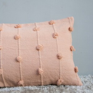 May include: A rectangular peach-colored decorative pillow with vertical rows of knotted yarn accents. The pillow is made of a textured fabric and rests on a light-colored, textured surface. The pillow is a soft, neutral color.