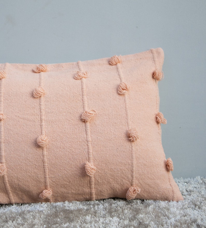 May include: A rectangular peach-colored decorative pillow with vertical rows of knotted yarn accents. The pillow is made of a textured fabric and rests on a light-colored, textured surface. The pillow is a soft, neutral color.
