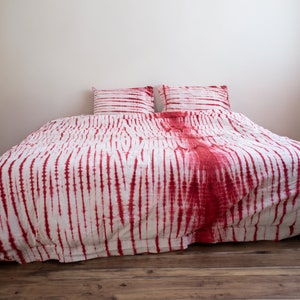 Może przedstawiać: Łóżko z białą i czerwoną pościelą w stylu tie-dye. Zestaw zawiera poszwę na kołdrę i dwie pasujące poszewki na poduszki. Czerwony barwnik jest w pionowe paski, tworząc odważny wygląd. Łóżko stoi na drewnianej podłodze.