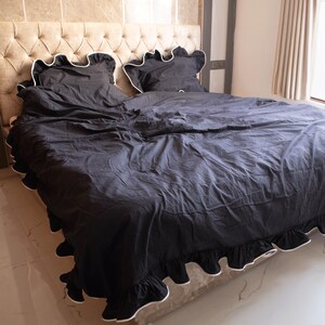 May include: Black bed linens with white trim, including a comforter and two pillowcases, are displayed on a bed. The bedding has a ruffled edge and is set against a beige upholstered headboard. The room has a neutral color palette.