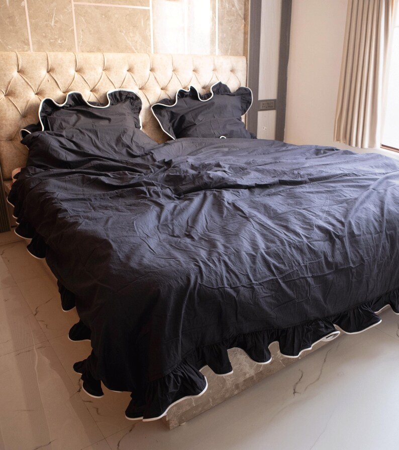 May include: Black bed linens with white trim, including a comforter and two pillowcases, are displayed on a bed. The bedding has a ruffled edge and is set against a beige upholstered headboard. The room has a neutral color palette.