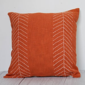 May include: Square orange decorative pillow with white stitched chevron pattern. The pillow has a textured fabric and a simple, modern design. The pillow is a square shape.