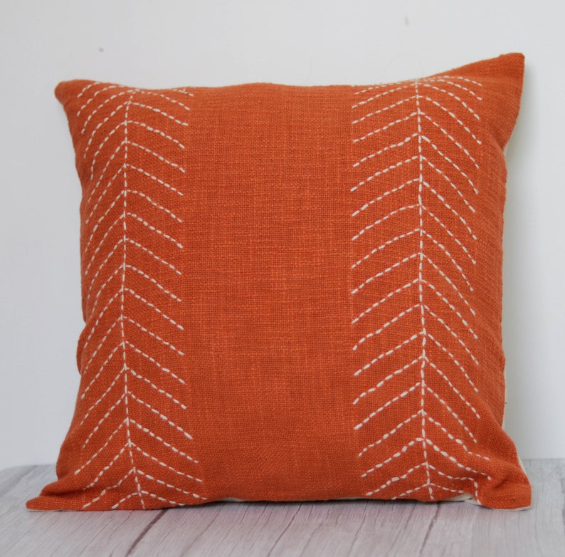 May include: Square orange decorative pillow with white stitched chevron pattern. The pillow has a textured fabric and a simple, modern design. The pillow is a square shape.