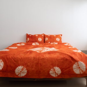 May include: An orange bedspread and two pillowcases with a white circular pattern. The bedding is on a wooden bed frame. The room has white walls and a window with natural light.