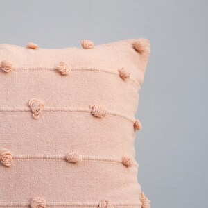 May include: A blush-colored decorative pillow with horizontal stitched lines and textured, knotted accents. The pillow has a square shape and is made of a woven fabric. The design adds a touch of texture and visual interest to the home decor.