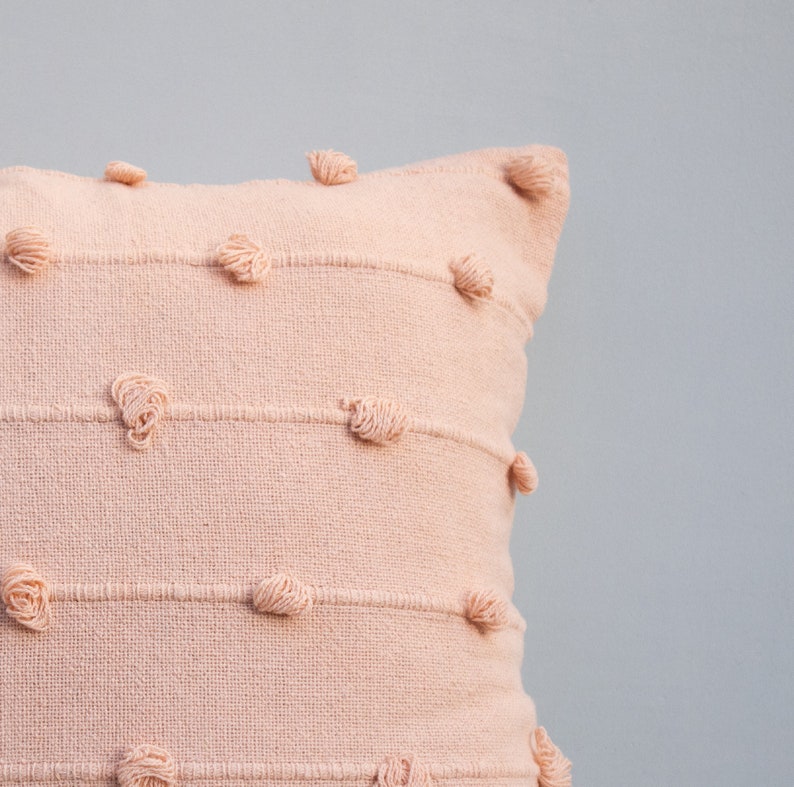 May include: A blush-colored decorative pillow with horizontal stitched lines and textured, knotted accents. The pillow has a square shape and is made of a woven fabric. The design adds a touch of texture and visual interest to the home decor.