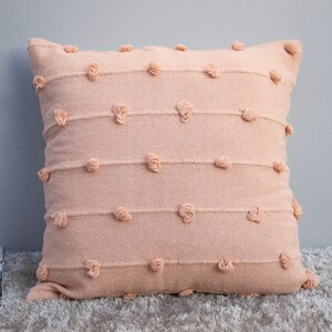 May include: A square peach-colored decorative pillow with horizontal rows of textured detailing and small, round, raised accents. The pillow is made of a soft, woven fabric and is placed on a light-colored rug.