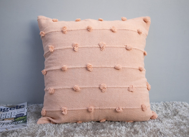 May include: A square peach-colored decorative pillow with horizontal rows of textured detailing and small, round, raised accents. The pillow is made of a soft, woven fabric and is placed on a light-colored rug.