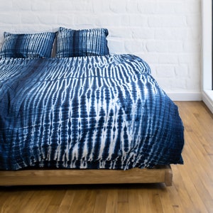 Puede incluir: Una cama con una funda nórdica y dos fundas de almohada a juego con estampado tie-dye azul y blanco. La ropa de cama está sobre una estructura de cama de madera, frente a una pared de ladrillo blanco y un suelo de madera. El estilo es moderno y bohemio.