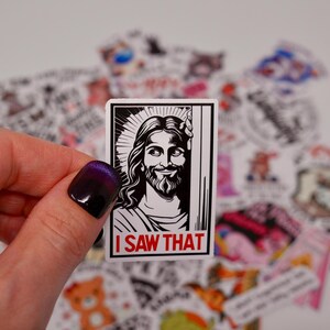 May include: A black and white sticker featuring a drawing of Jesus in a frame. The sticker has the text "I saw that" in red letters at the bottom.