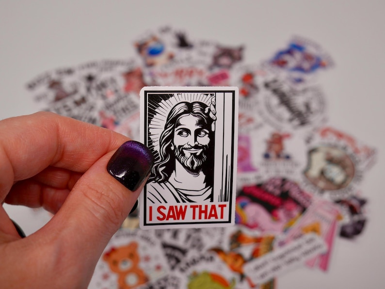 May include: A black and white sticker featuring a drawing of Jesus in a frame. The sticker has the text "I saw that" in red letters at the bottom.