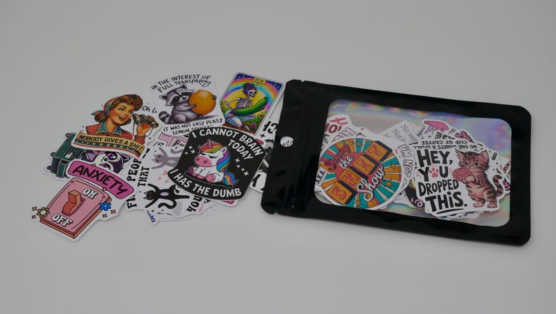 May include: A clear plastic bag filled with a variety of colourful stickers. The stickers feature humorous phrases and illustrations, including a raccoon, a unicorn, and a cat. The text on the stickers includes phrases like "I cannot brain today" and "Hey, you dropped this."