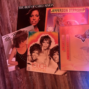 May include: A collection of vintage vinyl records featuring artists like Carly Simon, Jefferson Starship, Pat Benatar, and The Supremes. The records are displayed on a wooden surface.