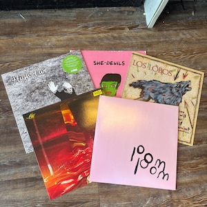 May include: A collection of vinyl records with various album covers. The covers feature diverse artwork, including text, illustrations, and abstract designs. The records are arranged on a wooden surface, showcasing a variety of colors and styles.