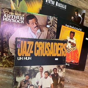 May include: A collection of five vinyl records, including albums by Arthur Prysock, Jazz Crusaders, Lonnie Liston Smith, Wynton Marsalis, and Sergio Mendes & Brasil '66. The records are stacked on a wooden surface.