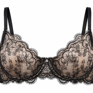 May include: A black and beige underwire bra featuring intricate black floral lace overlays on beige cups. The garment includes adjustable black shoulder straps, a scalloped lace edge along the neckline, and a small decorative crystal center detail.