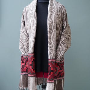 May include: A long, brown and white patterned cardigan with red and black geometric designs on the bottom. The cardigan has a fringe trim and is shown on a mannequin.