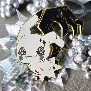 以下が含まれることがあります： 黒いアクセント、ダイヤモンド型のハイライト、様式化された顔を持つ漫画のようなキャラクターが特徴の白いエナメルピン。ピンには、金色のトリムと星空の背景が付いた黒い手が含まれています。ピンは銀色と灰色の背景に配置されています。