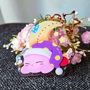May include: A pink and purple character with a yellow star on its head is sleeping on a yellow crescent moon. The character is wearing a purple and yellow hat. The crescent moon is attached to a gold chain with a star charm.