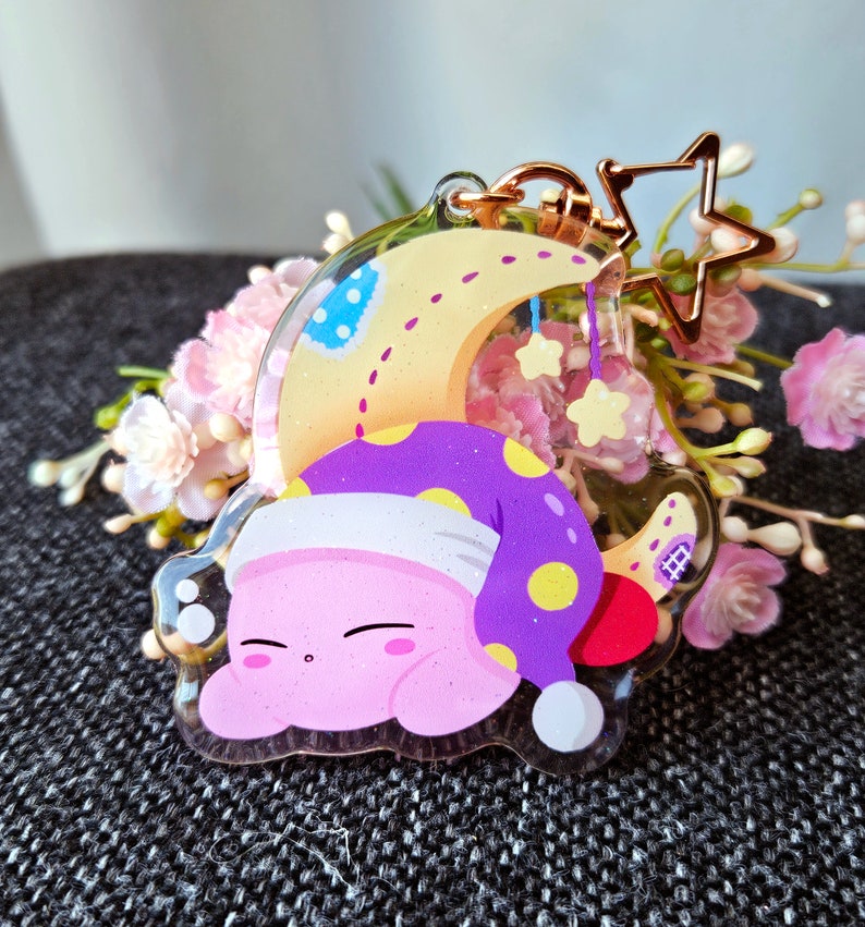 May include: A pink and purple character with a yellow star on its head is sleeping on a yellow crescent moon. The character is wearing a purple and yellow hat. The crescent moon is attached to a gold chain with a star charm.