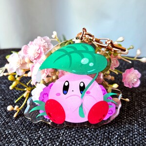 May include: A pink and green character keychain with a leaf-shaped canopy. The character has large eyes and a pink body with red accents.
