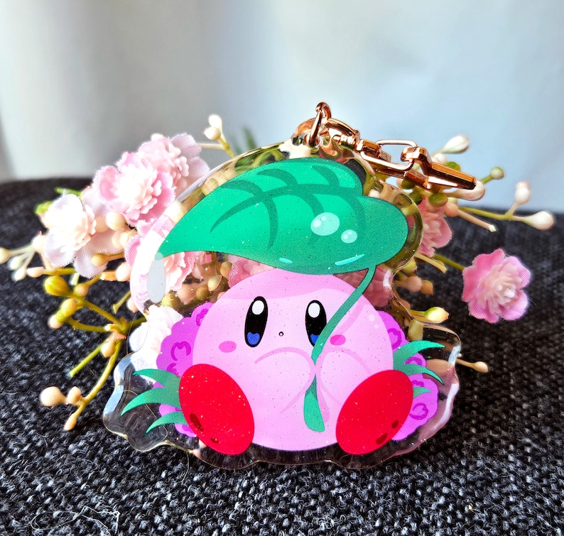 May include: A pink and green character keychain with a leaf-shaped canopy. The character has large eyes and a pink body with red accents.
