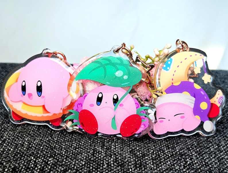 May include: Four pink and white cartoon character keychains with different designs. The first keychain features a pink character with a yellow and orange pastry. The second keychain features a pink character with a green leaf. The third keychain features a pink character with a purple hat and a yellow moon. The fourth keychain features a pink character with a yellow star.