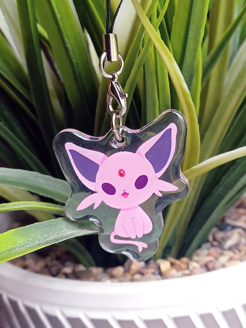 May include: A clear plastic charm with a pink and purple cartoon character. The character has a pink body, purple ears, and a pink gem on its forehead. The charm is attached to a black string.