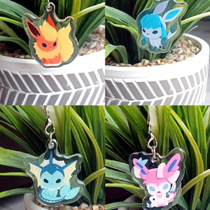 May include: Four clear acrylic charms featuring different Pokemon characters. The charms are attached to silver chains. The charms are displayed against a background of green plants in white pots.